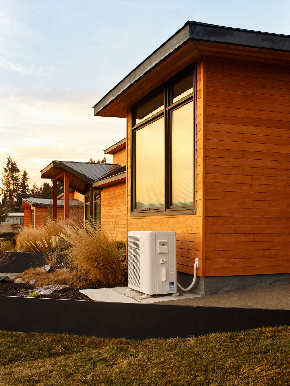 A cedar-clad modern home at golden hour with a new heat pump on a stone pad beside the wall